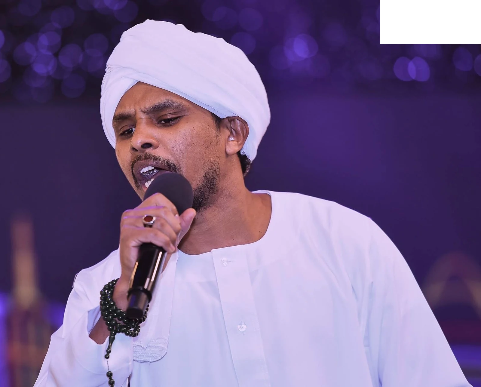 Ahmed Fathalla performing in a white jalabiya and turban, holding a microphone